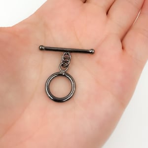May include: A gunmetal-colored toggle clasp, featuring a circular ring connected to a bar with a series of small rings. The clasp is designed for jewelry making and is shown against a neutral background.