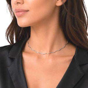 May include: A delicate silver-toned necklace with a wavy design. The necklace is worn on a person wearing a black satin blazer. The necklace appears to be made of small, connected beads or links, creating a subtle, shimmering effect.