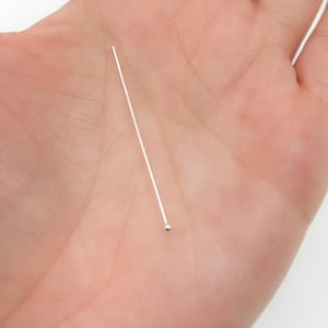 Könnte beinhalten: Ein einzelner silberfarbener Kopfstift mit einem kleinen Kugelende, der auf einem hellen, hautfarbenen Hintergrund liegt. Der Stift ist gerade und dünn, mit einer glatten, reflektierenden Oberfläche. Das Kugelende ist eine kleine, runde Form.