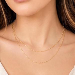 Puede incluir: Un collar de cadena doble de oro con eslabones ovalados alargados y delicados. Las dos hebras del collar descansan a longitudes ligeramente diferentes alrededor del cuello, creando una apariencia superpuesta con un acabado metálico pulido.