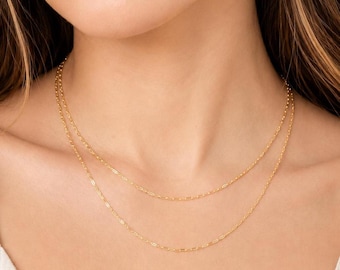 May include: A double-layered gold chain necklace featuring delicate, elongated oval links. The two strands of the necklace rest at slightly different lengths around the neck, creating a layered appearance with a polished, reflective metallic finish.