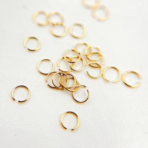 May include: A collection of small, open gold-colored rings scattered on a white surface. The rings are circular with a small gap, suggesting they are designed to be opened and closed. The lighting highlights the metallic sheen of the jewelry.
