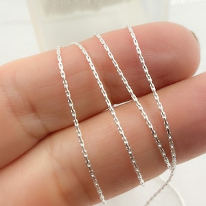 May include: Close-up of a silver chain, showing the delicate, twisted design of the links. The chain is laid out in a few layers on a finger, highlighting its thinness.