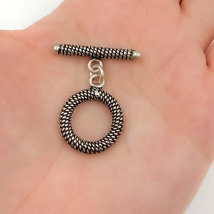 May include: A silver-toned toggle clasp, featuring a textured, cylindrical bar and a circular ring. The ring and bar are connected by a small chain. The clasp is designed for jewelry making and is shown against a neutral background.