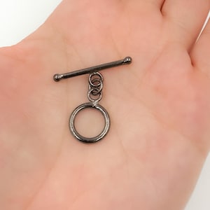 May include: A dark gray toggle clasp, featuring a straight bar with rounded ends and a circular ring. The bar is connected to the ring via a series of small, interlocking loops. This jewelry finding is designed for use in necklaces and bracelets.