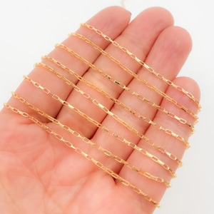 Puede incluir: Un delicado collar de cadena de color dorado con un diseño de eslabones rectangulares. La cadena se sostiene en una mano, mostrando sus finos detalles y la calidad cálida y reflectante del metal. Los eslabones son pequeños y espaciados uniformemente.