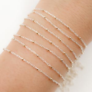 Puede incluir: Un conjunto de pulseras de cadena plateadas con cuentas doradas. Las pulseras están superpuestas en la muñeca de una persona.