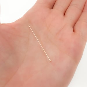 May include: A single, thin, gold-colored pin, likely for jewelry making or crafts, is held in a person's open palm. The pin is straight and has a small, rounded head. The background is a light skin tone.