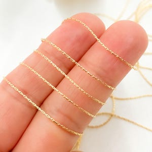 May include: Close-up of delicate gold-colored chain. The chain is thin and has a textured, faceted appearance. The chain is draped across two fingers, showcasing its flexibility and design. The background is a soft, neutral color.