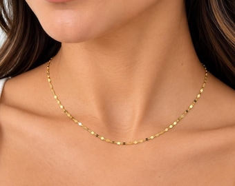 Puede incluir: Un delicado collar de cadena de oro con pequeños eslabones ovalados planos y reflectantes que captan la luz. El collar descansa sobre el cuello, mostrando un acabado metálico brillante con un patrón consistente y repetitivo de eslabones.