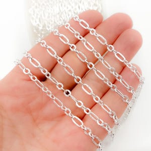 May include: Close-up of a silver-colored chain with oval and elongated links. The chain is held in a hand, showcasing its texture and design. The links have a textured, glittery appearance, reflecting light.