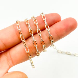 Op de afbeelding: Een close-up van een zilveren en gouden ketting. De ketting heeft afwisselende ovale en gedraaide schakels. De ketting wordt gepresenteerd tegen een lichte achtergrond.