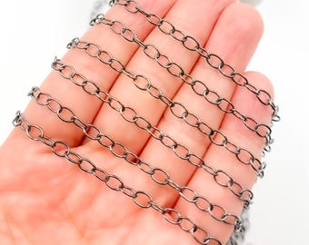 Op de afbeelding: Close-up van een lengte donkergrijze ovale schakelketting. De ketting wordt in een hand gehouden tegen een witte achtergrond. De schakels zijn uniform van grootte en vorm, waardoor een doorlopend patroon ontstaat.