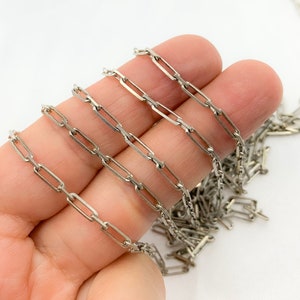 May include: A silver-colored chain with a paperclip link design. The chain is held in a hand against a white background. The links are rectangular and connected end-to-end.