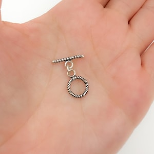 May include: Silver toggle clasp with a twisted rope design. The clasp has a round ring and a bar that slides through the ring.