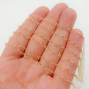 Könnte beinhalten: Eine zarte goldfarbene Kette mit einem sich wiederholenden Herzmuster. Die Kette wird in einer Hand vor einem weißen Hintergrund gehalten. Die Herzen sind klein und gleichmäßig verteilt, was ein romantisches und elegantes Design ergibt, das sich für die Schmuckherstellung eignet.