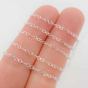 Könnte beinhalten: Nahaufnahme von mehreren Strängen einer filigranen, silberfarbenen Kette. Jedes Glied ist wie ein Unendlichkeitssymbol geformt. Die Ketten werden vor einem hellen Hintergrund präsentiert, der ihr kompliziertes Design und ihren metallischen Glanz hervorhebt. Ideal für die Schmuckherstellung.