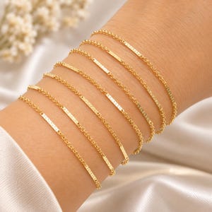 Könnte beinhalten: Ein Set aus sieben goldfarbenen Kettenarmbändern, die jeweils ein schmales, rechteckiges Element mit strukturierter Oberfläche in der Mitte aufweisen. Die Armbänder werden zusammen getragen und zeigen die feinen Gliederketten und das einheitliche metallische Finish.