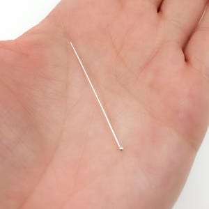 Könnte beinhalten: Eine einzelne, dünne, silberfarbene Stecknadel mit einem kleinen Kugelkopf. Die Nadel wird in der offenen Handfläche einer Person gehalten, wodurch ihr filigranes Design und ihr metallischer Glanz zur Geltung kommen. Die Nadel ist zum Nähen oder Basteln bestimmt.