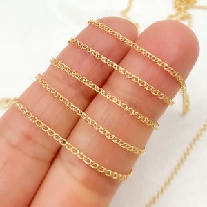 May include: Close-up of a delicate gold chain with a wheat-style design. The chain is draped across fingers, showcasing its intricate links and warm, golden hue. The background is a soft, neutral color, highlighting the chain's details.