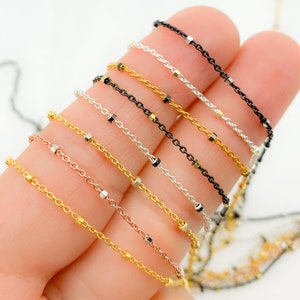 May include: A close-up of a hand holding a variety of beaded chains in different colors, including gold, silver, rose gold, and black.