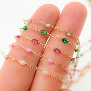 Könnte beinhalten: Nahaufnahme von mehreren Strängen zarter Goldketten mit farbigen, oval geformten Perlen. Die Perlen sind in Rosa-, Grün- und Klartönen gehalten und bilden ein lebendiges Design. Die Ketten werden zwischen zwei Fingern gehalten.