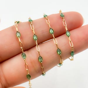 Puede incluir: Una cadena dorada con pequeñas cuentas de vidrio verde de forma ovalada y un patrón texturizado en forma de estrella. Las cuentas están espaciadas uniformemente entre eslabones metálicos dorados alargados, creando un diseño decorativo repetitivo.
