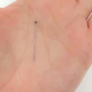 Könnte beinhalten: Eine einzelne silberfarbene Stecknadel mit einem kleinen, runden, dunkelgrauen Kopf. Die Nadel ist gerade und schlank, für Bastel- oder Nähprojekte geeignet. Die Nadel wird in einer Hand gehalten.