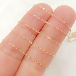 以下が含まれることがあります： 繊細なゴールドカラーのチェーンのクローズアップ。チェーンは小さな楕円形のリンクで構成されています。チェーンは2本の指の間に保持され、背景は白です。