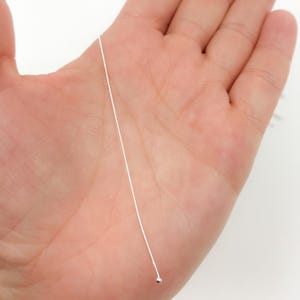 Könnte beinhalten: Ein dünner, silberfarbener Faden mit einer kleinen, runden Perle am Ende. Der Faden wird in einer offenen Hand vor einem weißen Hintergrund gehalten. Die Perle scheint metallisch zu sein und reflektiert Licht.