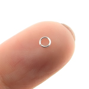 May include: A small, silver-colored jump ring, open at one point, is shown against a blurred background. The ring is circular and appears to be made of metal. It is likely used for jewelry making or crafts.
