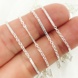 May include: Close-up of several delicate silver necklaces. Each necklace features a fine chain with small, interconnected links. Some necklaces have textured bar pendants, while others have plain bars. The necklaces are displayed against a light background.