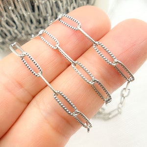 May include: Close-up of a silver-colored chain with oval links. Each link has a textured, patterned surface. The chain is held in a hand, with a blurred background of more chain.