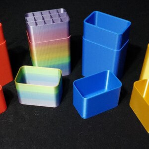May include: A set of six 3D printed storage containers in red, blue, yellow, and rainbow colors. The containers are stacked and unstacked, showing the different sizes and shapes.