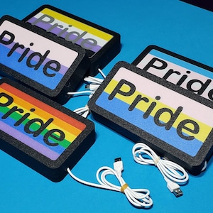 May include: Rectangular black boxes with the word "Pride" in bold, white letters. The boxes feature various Pride flag designs, including rainbow, transgender, and non-binary flags. White USB cables are also visible.