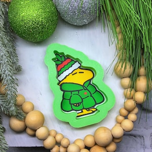 May include: A green, Christmas-themed mold featuring a cartoon character in a winter hat and coat. The character is yellow with a green coat and hat with red and white stripes. The mold is surrounded by Christmas ornaments and decorative elements.