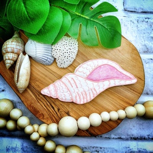 Puede incluir: Una galleta de azúcar rosa y blanca con forma de concha marina con un interior rosa. La galleta está sobre una tabla de madera con otras conchas marinas y una hebra de cuentas de madera.