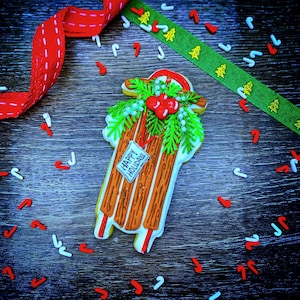 May include: A festive Christmas cookie shaped like a wooden sled, decorated with green frosting, red berries, and a tag that reads "Happy Holidays." Red and white candy canes and ribbons surround the sled on a wooden surface.