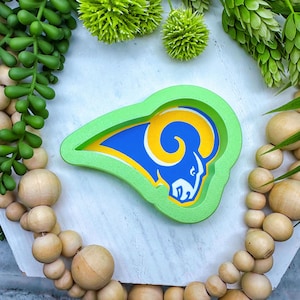 May include: A green and blue Los Angeles Rams logo, featuring a ram's head, is displayed on a white surface. The logo is surrounded by a green border. The image is decorated with green plants and wooden beads.