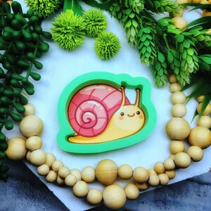 May include: A green plastic cookie cutter shaped like a cartoon snail with a pink shell and a yellow body. The snail has a smiling face.