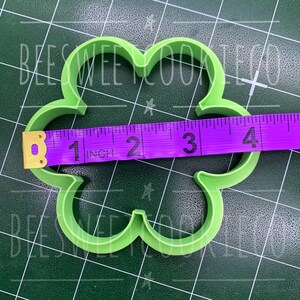 Pu&ograve; includere: Un tagliabiscotti verde chiaro a forma di quadrifoglio. Il metro a nastro viola mostra che il tagliabiscotti &egrave; largo circa 10 cm. Lo sfondo &egrave; un tappetino da taglio verde scuro con il testo "BEESWEE COOKIE CO".