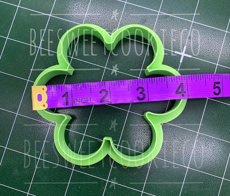 Pu&ograve; includere: Un tagliabiscotti verde chiaro a forma di quadrifoglio. Il metro a nastro viola mostra che il tagliabiscotti &egrave; largo circa 10 cm. Lo sfondo &egrave; un tappetino da taglio verde scuro con il testo "BEESWEE COOKIE CO".