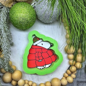 May include: A Snoopy-shaped Christmas ornament in a red coat and hat, outlined in green. The ornament is surrounded by Christmas decorations, including green and silver glittery ornaments, pine needles, and wooden beads.