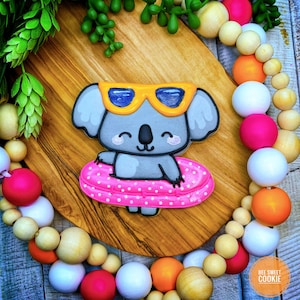 May include: A gray koala cookie wearing yellow sunglasses and floating on a pink and white polka dot inner tube. The cookie is on a wooden surface with a beaded garland in the background.