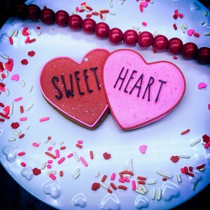 Pode incluir: Dois biscoitos em forma de coração, um rosa e outro vermelho, decorados com glacê branco e as palavras "Sweet" e "Heart". Os biscoitos estão em um prato branco com granulado vermelho e branco.