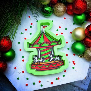 May include: Green, red, and white cookie cutter shaped like a carousel with horses. The cutter is on a white surface with red and green sprinkles.