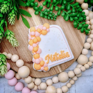 May include: A white cookie decorated with a border of orange and pink balloons and the name "Natalia" in orange icing.