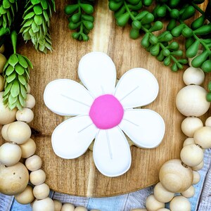 Pu&ograve; includere: Un biscotto a forma di margherita bianca con un centro rosa acceso. Il biscotto &egrave; su una superficie di legno, circondato da piante verdi e perline di legno. Il biscotto a forma di fiore ha petali bianchi e un centro rosa.