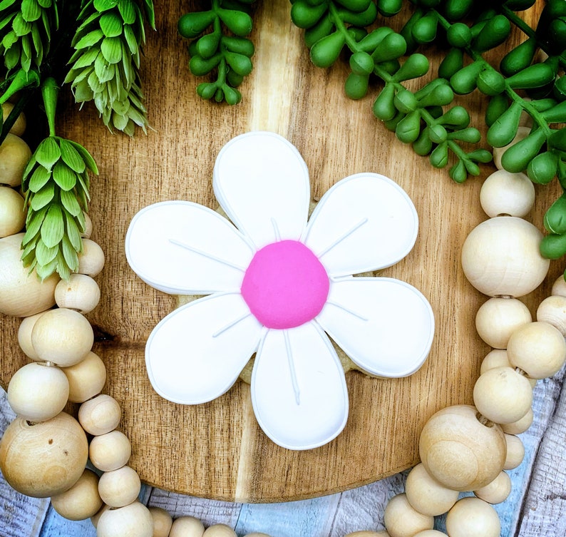 Pu&ograve; includere: Un biscotto a forma di margherita bianca con un centro rosa acceso. Il biscotto &egrave; su una superficie di legno, circondato da piante verdi e perline di legno. Il biscotto a forma di fiore ha petali bianchi e un centro rosa.