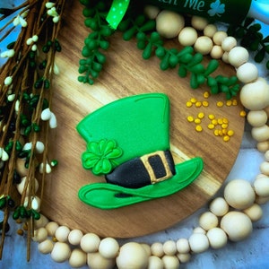 May include: Green and gold leprechaun hat cookie decorated with a shamrock. The cookie is on a wooden surface with gold sprinkles and wooden beads.